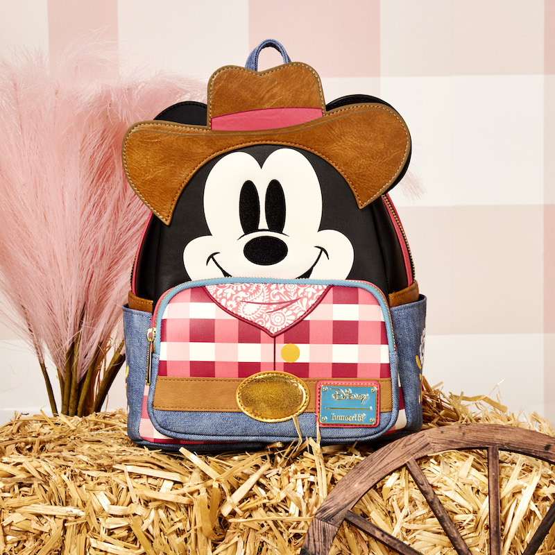 Loungefly Western Mickey Mouse Mini Backpack sitting on a hay bale with pink feathers behind it and sitting against a pink and white gingham background. 